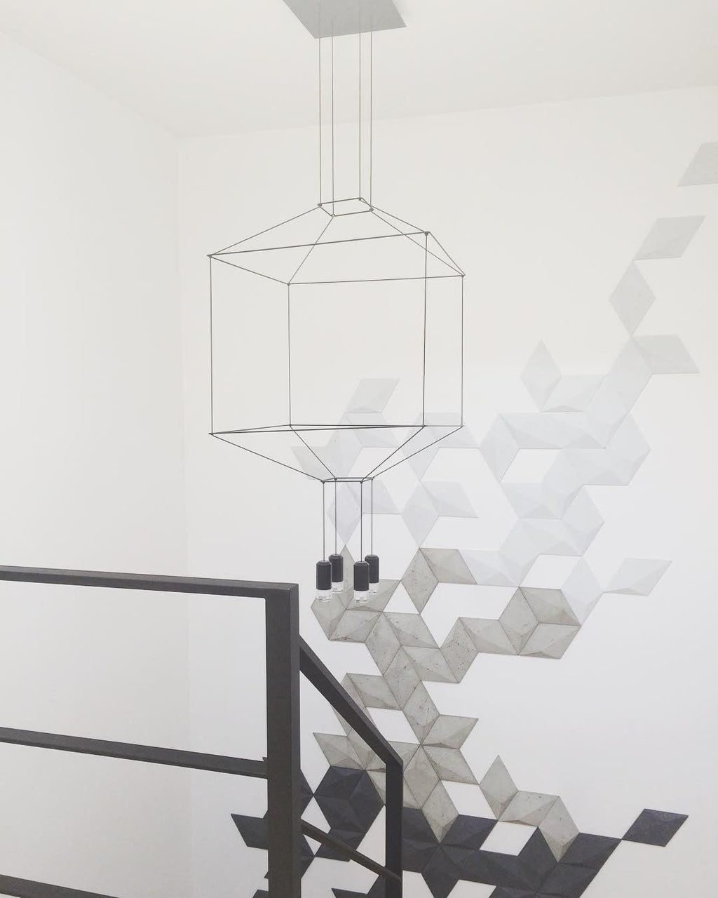 Square wireframe pendant hung in a stairwell beside a geometric tiled wall and a black railing, from Clérachic.