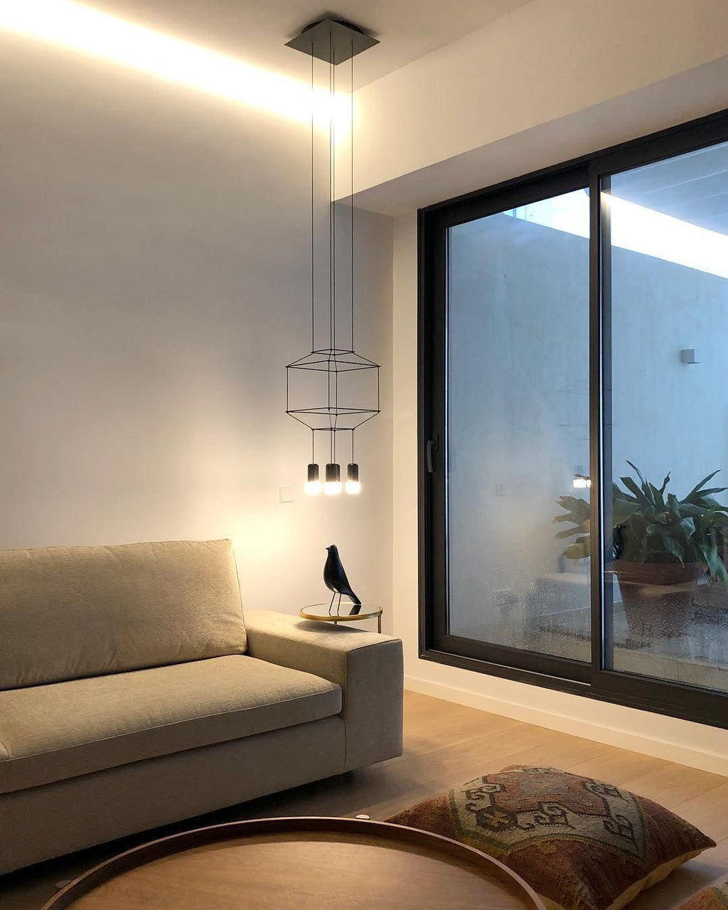 Small wireframe pendant in a living room with a sofa and a glass sliding door, with a cushion on the floor, from Clérachic.