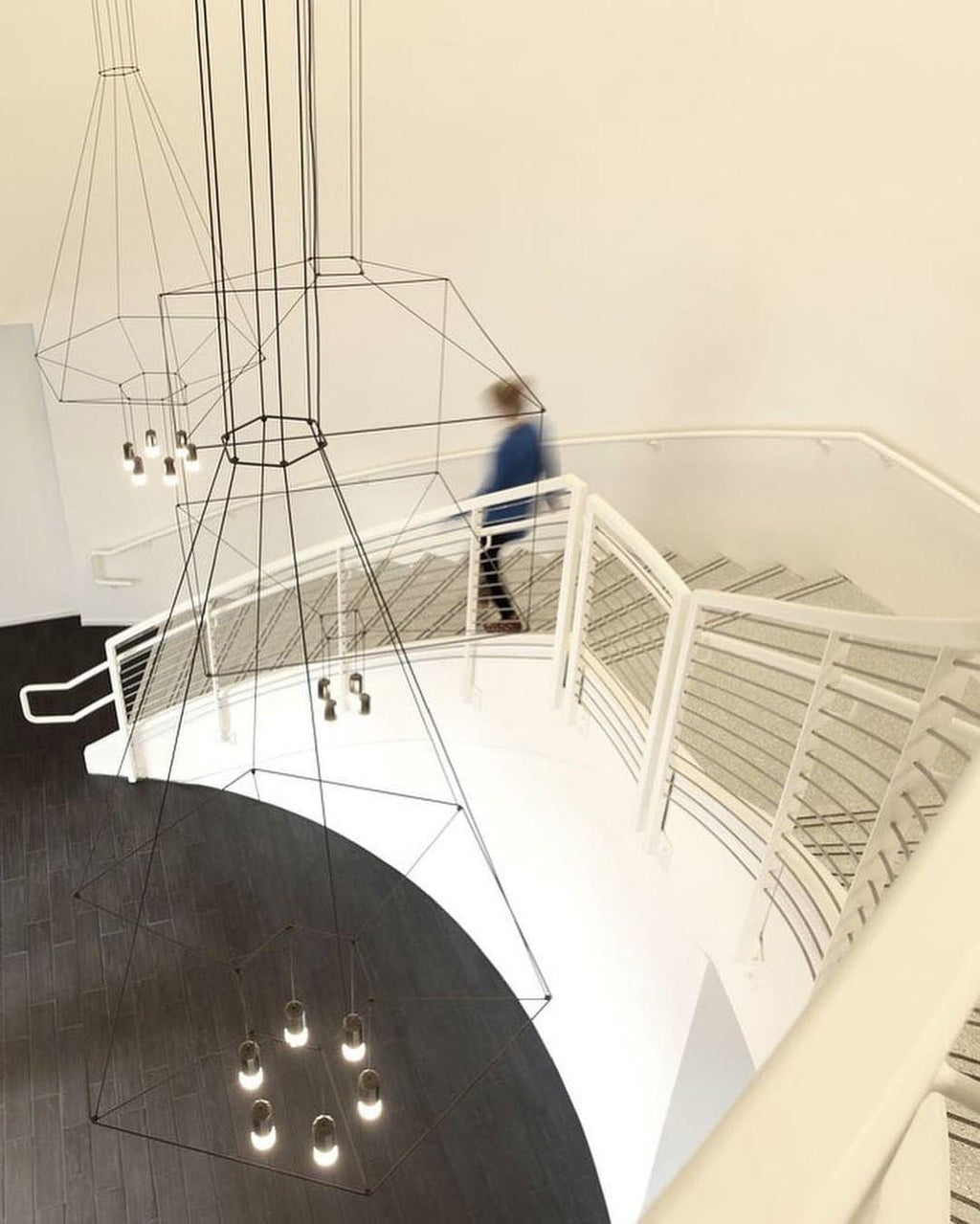 Wireframe pendant seen from above a white spiral staircase, its lights hanging down the stairwell, from Clérachic.