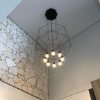 Octagonal wireframe pendant with a black canopy and lit bulbs, below a white ceiling beside a stone wall, from Clérachic.