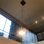 Cube-framed pendant with glowing bulbs, seen from below beside a glass balustrade and a high-rise tower, from Clérachic.