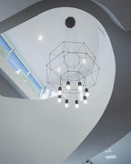 Black wireframe pendant with a canopy and glowing lights, seen from below in a curved white stairwell, from Clérachic.