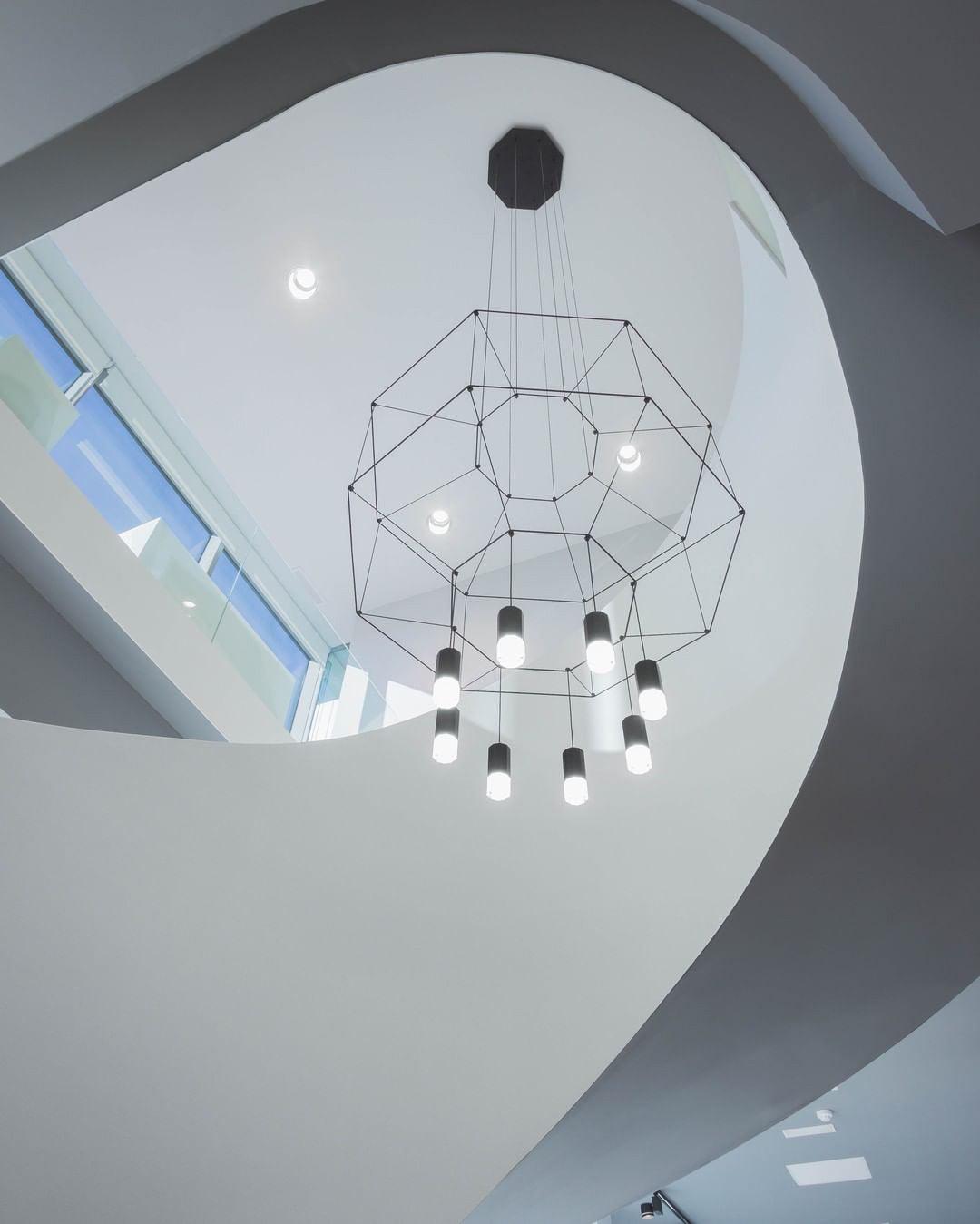 Black wireframe pendant with a canopy and glowing lights, seen from below in a curved white stairwell, from Clérachic.