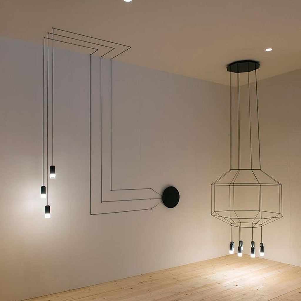 Black wireframe pendant with a round canopy beside a cluster of lights fed by lines of cable on the wall, from Clérachic.