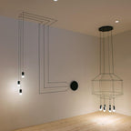 Black wireframe pendant with a round canopy beside a cluster of lights fed by lines of cable on the wall, from Clérachic.