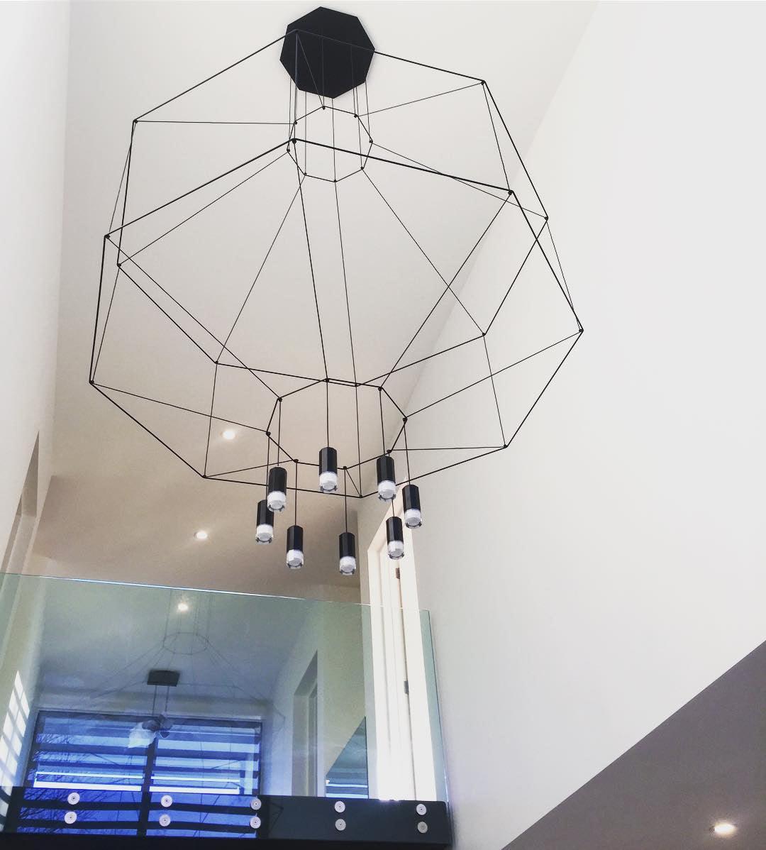Looking up at an octagonal wireframe pendant on a black canopy, its lights hanging below, in a tall stairwell, from Clérachic.