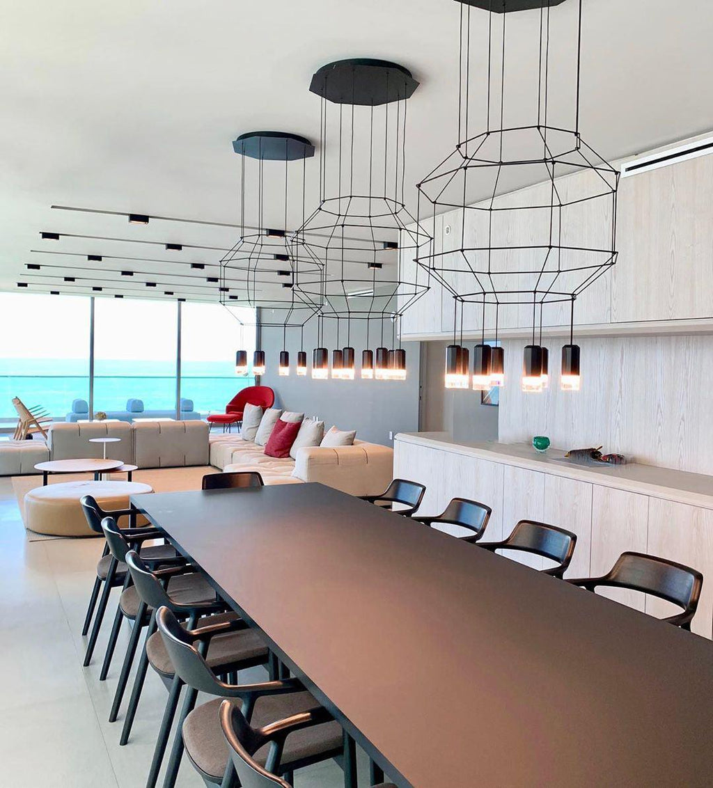 Three octagonal wireframe pendants in a row above a long dark table with chairs, in a room by the sea, from Clérachic.