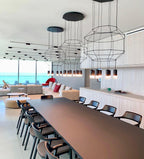 Three octagonal wireframe pendants in a row above a long dark table with chairs, in a room by the sea, from Clérachic.