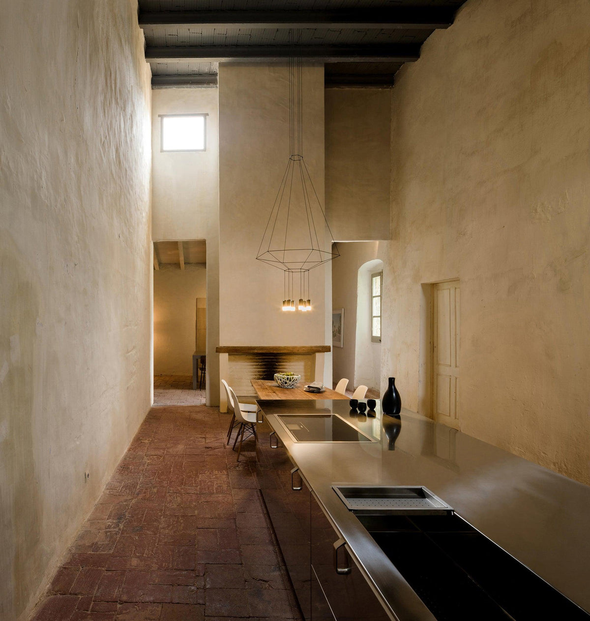 Cone-shaped wireframe pendant at the end of a long kitchen with plaster walls and a brick floor, from Clérachic.