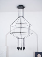 Black wireframe pendant on a ceiling canopy against a white wall, with its tiered cage and unlit light heads, from Clérachic.