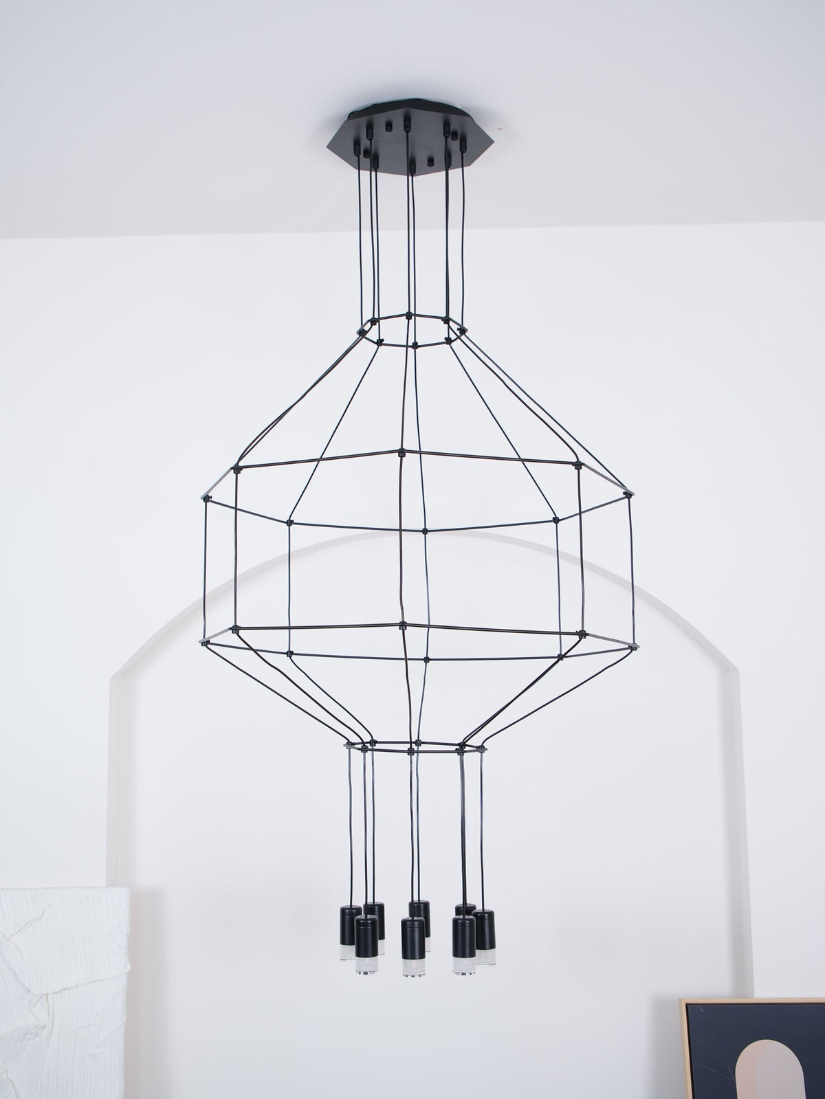 Black wireframe pendant on a ceiling canopy against a white wall, with its tiered cage and unlit light heads, from Clérachic.