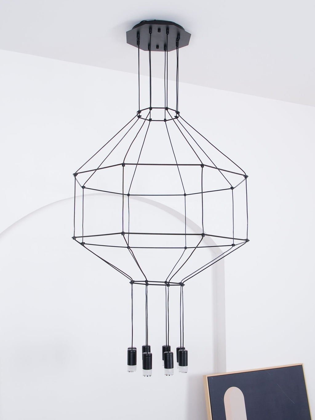 Unlit Clérachic wireframe pendant with a tiered cable cage and small black light heads, hung before a white arched wall.