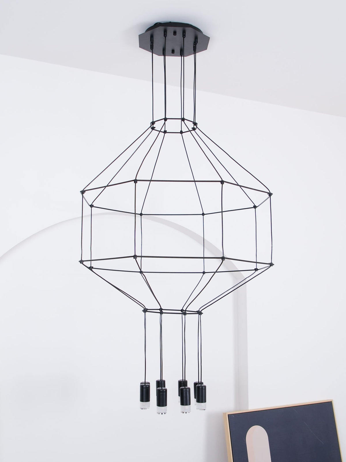 Unlit Clérachic wireframe pendant with a tiered cable cage and small black light heads, hung before a white arched wall.