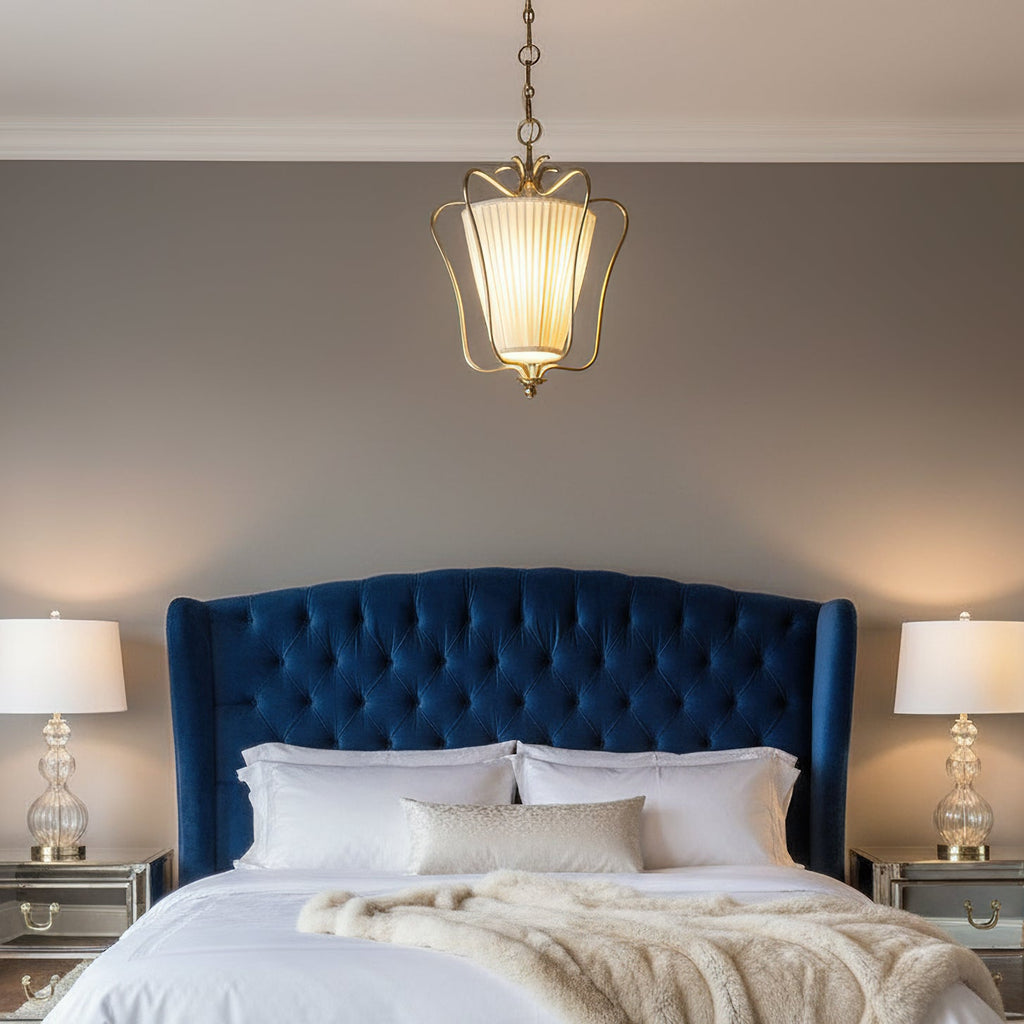 Lit Clérachic brass pendant in a bedroom with a blue tufted headboard, two glass table lamps and a cream throw.