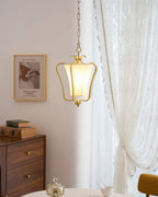 Clérachic brass pendant with a twisted-pleat shade, lit beside a lace curtain and a framed print above a walnut chest of drawers.