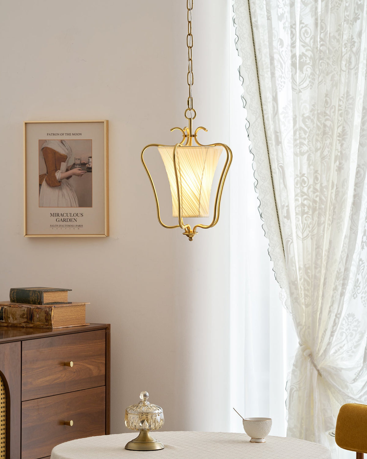 Lit Clérachic pendant with a diagonally pleated shade, a lace curtain behind it and a glass dish on a round table below.