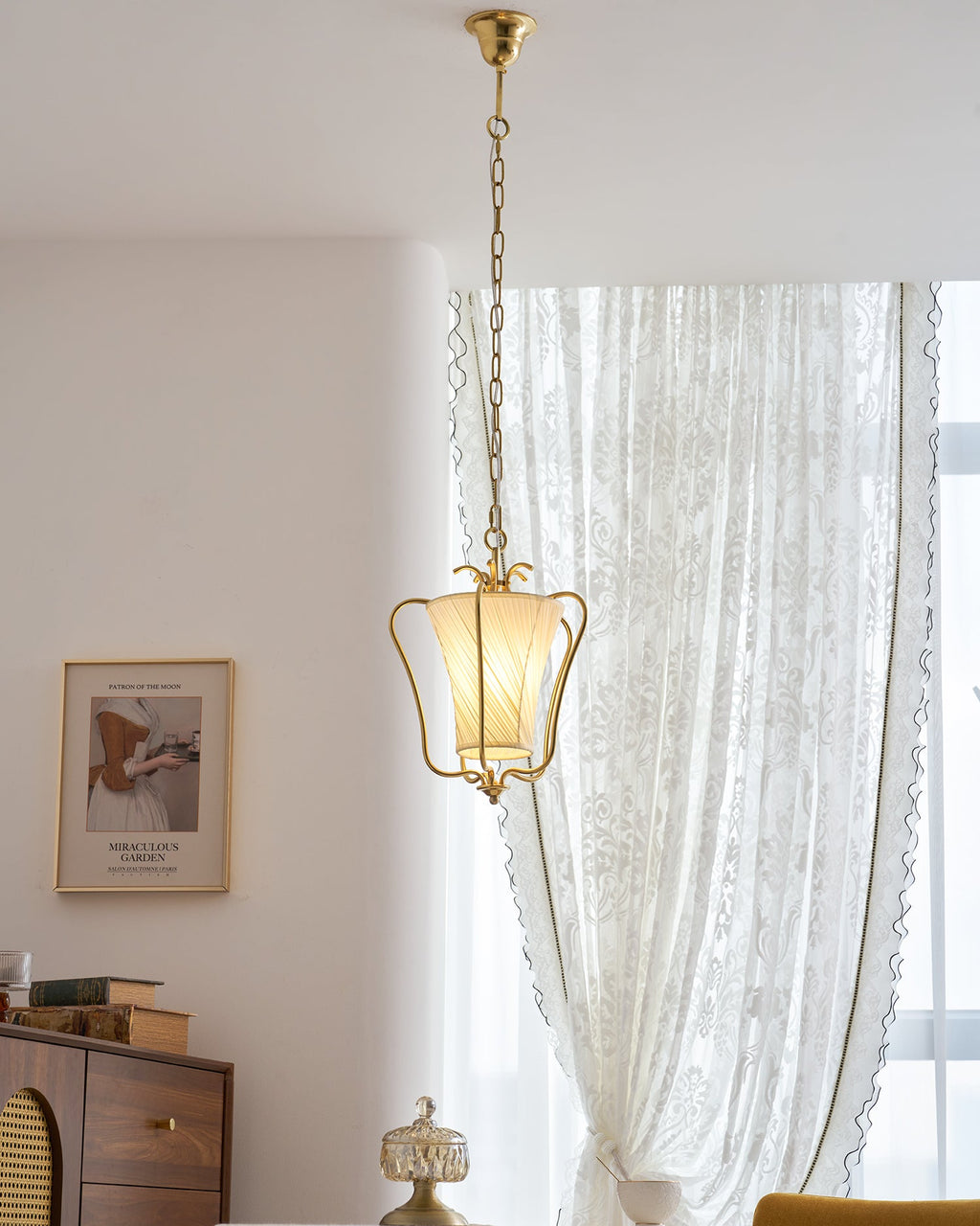 Tall view of this Clérachic brass pendant on its long chain, with a lace curtain, a framed print and a wooden cabinet beyond.