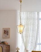 Tall view of this Clérachic brass pendant on its long chain, with a lace curtain, a framed print and a wooden cabinet beyond.