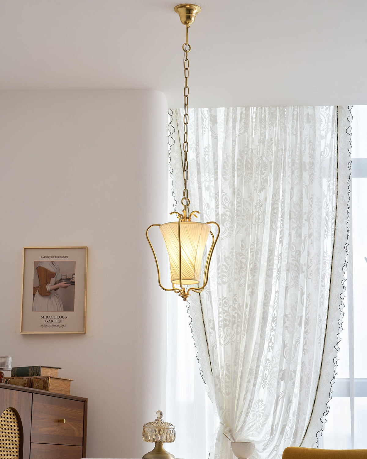 Tall view of this Clérachic brass pendant on its long chain, with a lace curtain, a framed print and a wooden cabinet beyond.