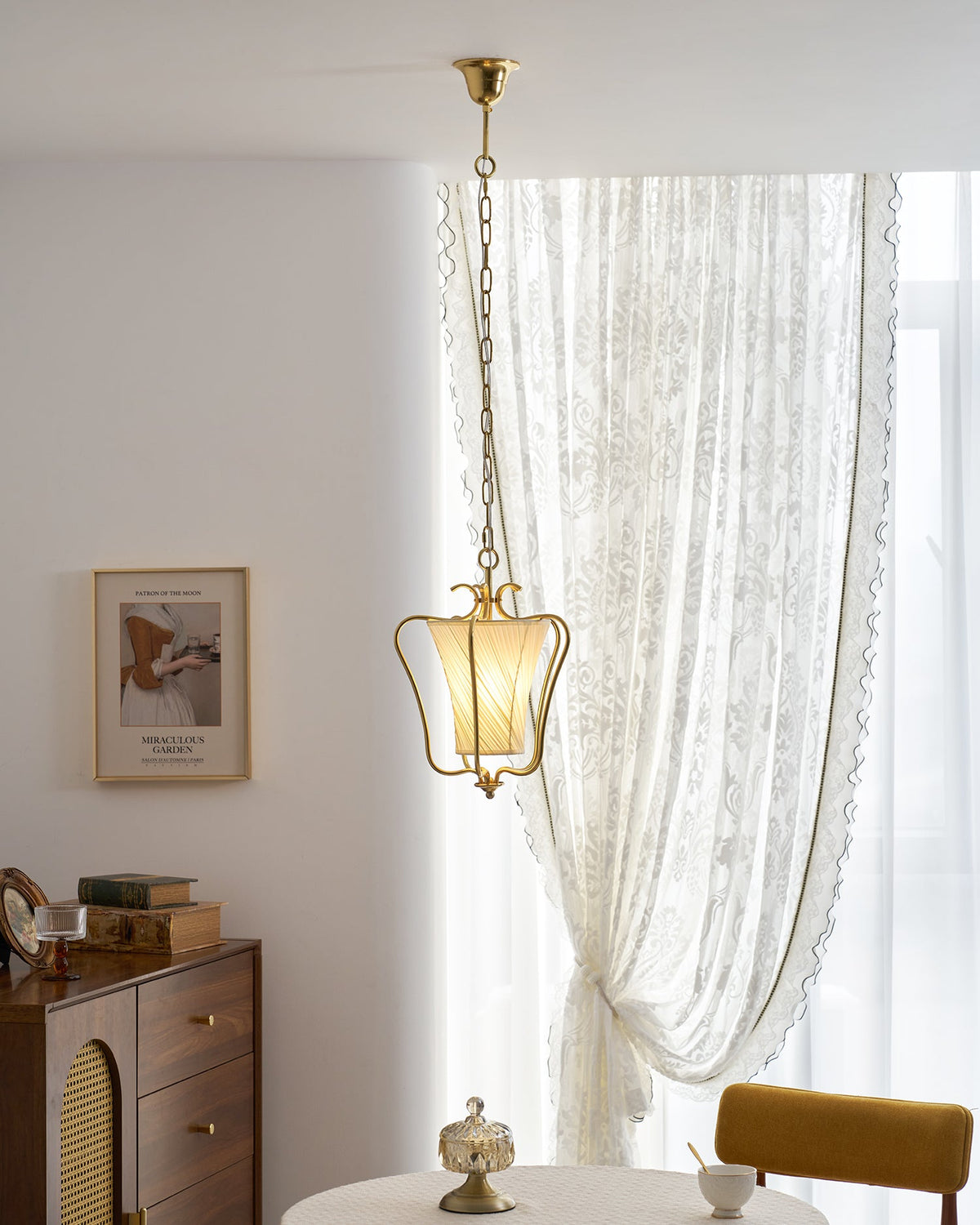 Clérachic pendant with a diagonal-pleat shade beside a sheer curtain, with a walnut sideboard and a framed print below.