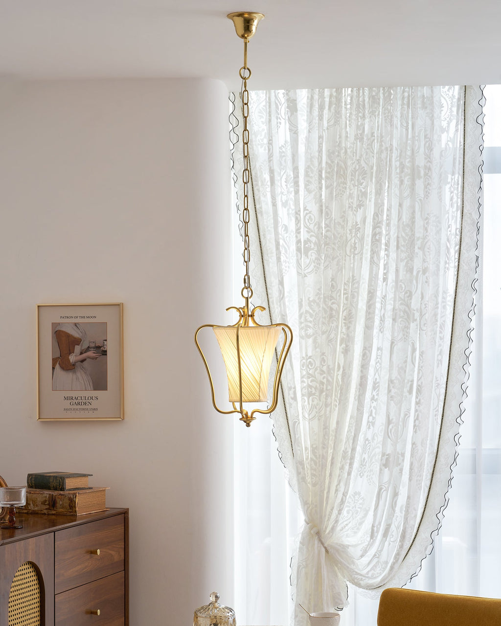 Clérachic brass lantern pendant with a twisted fabric shade in front of a lace curtain, with a small glass dish at the bottom.