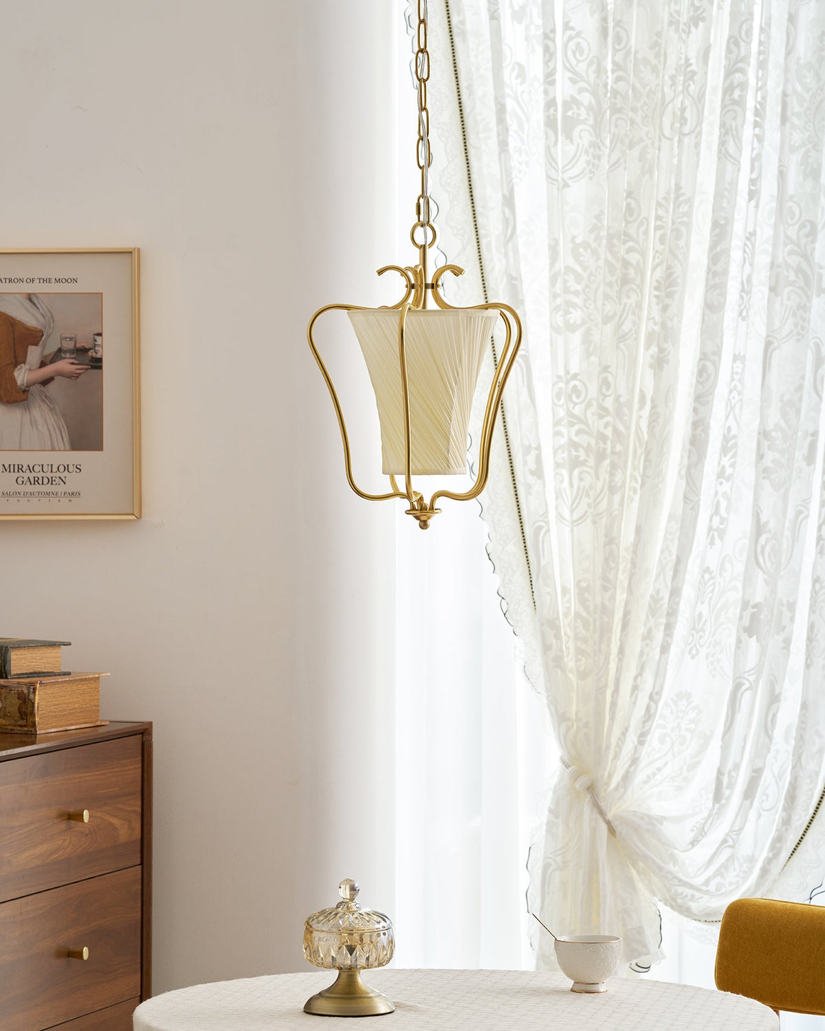 Unlit Clérachic pendant with a cream diagonal-pleat shade, hanging beside a framed print and a lace curtain.