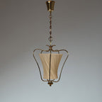 Clérachic brass pendant with a cream diagonally pleated shade, hung against a grey studio backdrop with a brass canopy.
