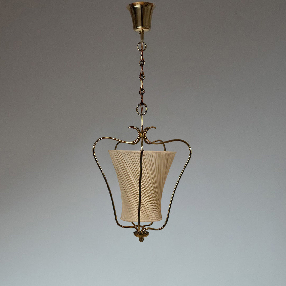 Clérachic brass pendant with a cream diagonally pleated shade, hung against a grey studio backdrop with a brass canopy.