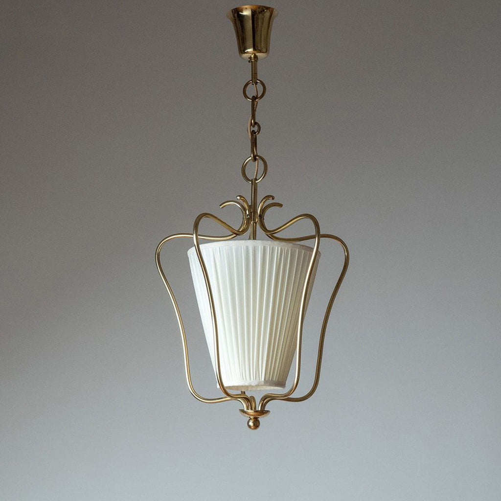 Clérachic brass pendant with a white vertically pleated shade and a curled crown, hung against a grey studio backdrop.