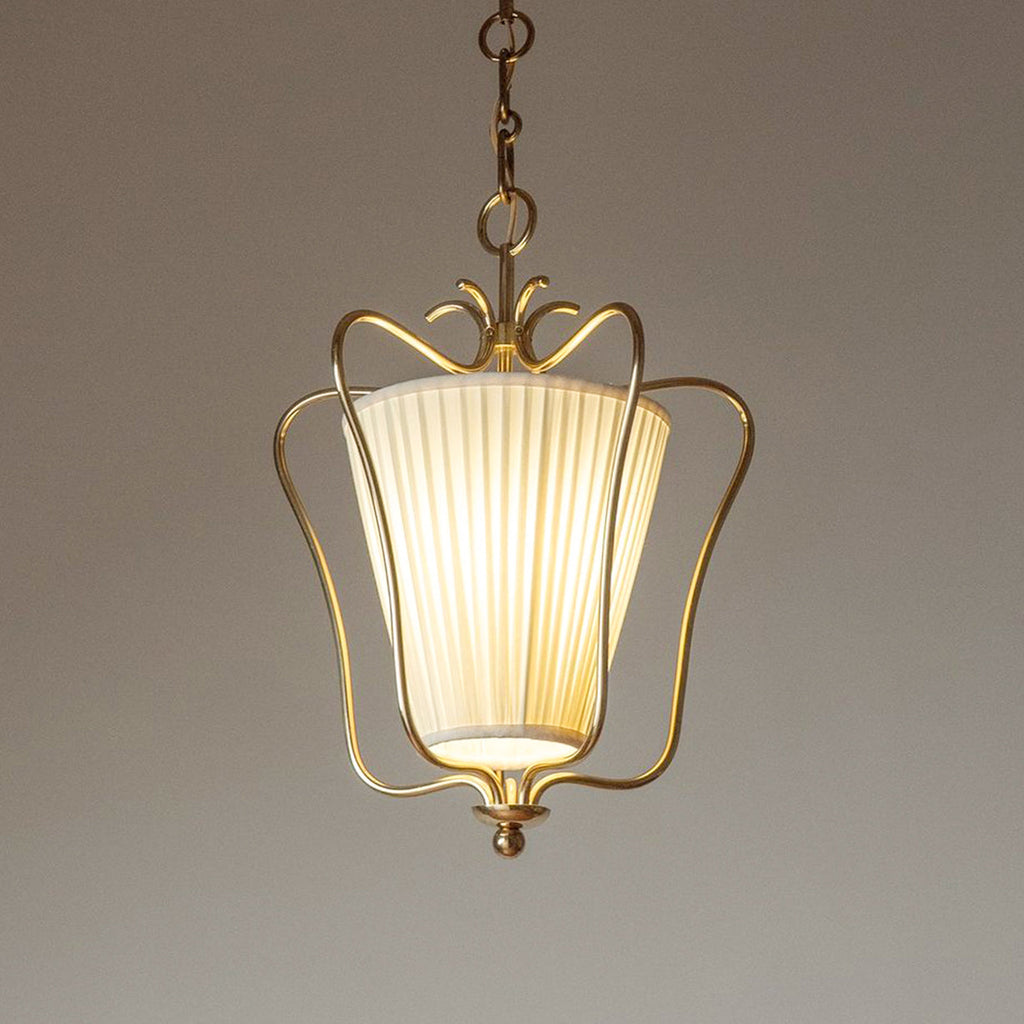 Glowing Clérachic pendant with a vertically pleated shade inside the brass frame, against a warm taupe backdrop.