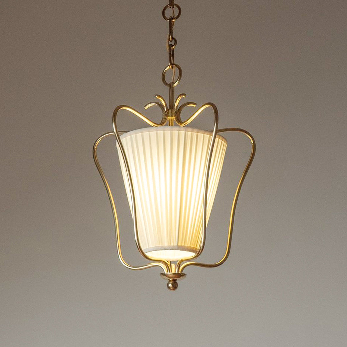 Glowing Clérachic pendant with a vertically pleated shade inside the brass frame, against a warm taupe backdrop.