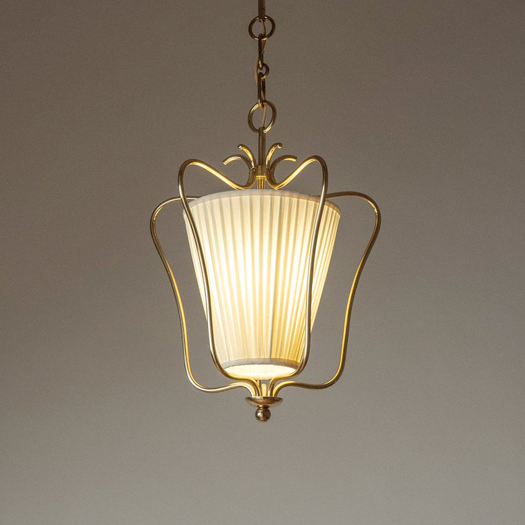 Clérachic pendant with a pleated shade lit from inside, its brass crown and drop finial in focus, on a taupe backdrop.