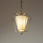Clérachic pendant with a pleated shade lit from inside, its brass crown and drop finial in focus, on a taupe backdrop.
