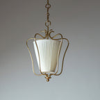 Unlit Clérachic pendant with a white vertically pleated shade in a brass frame, on a pale blue-grey backdrop.