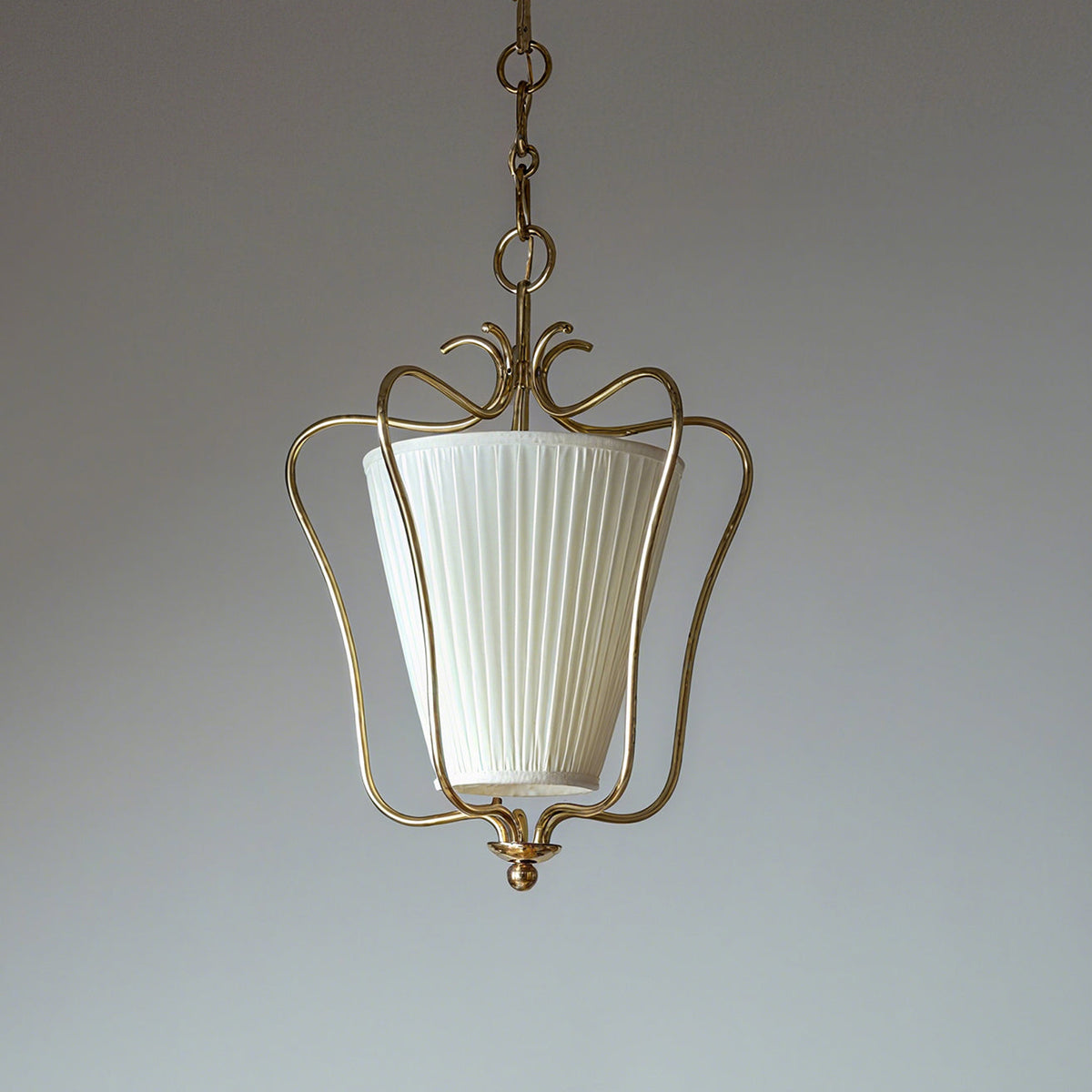 Unlit Clérachic pendant with a white vertically pleated shade in a brass frame, on a pale blue-grey backdrop.