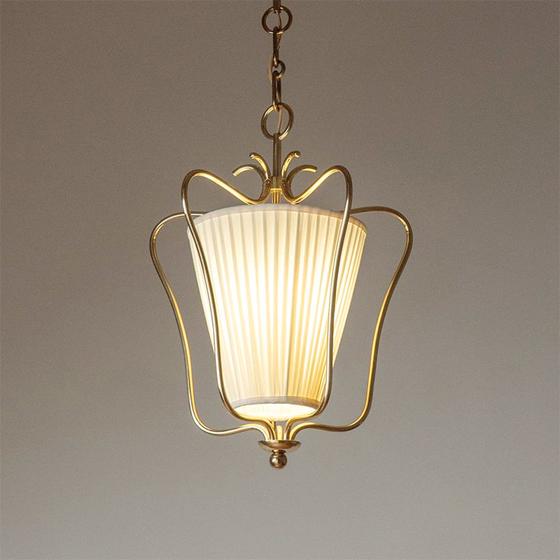 Close view of a lit Clérachic brass pendant with a vertically pleated shade and a curled crown, on a taupe backdrop.