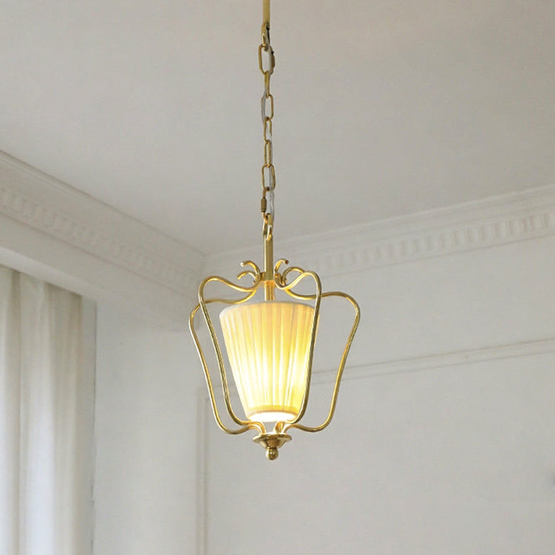 Lit Clérachic brass pendant with a pleated shade, hanging from a chain beside a white cornice and a curtain edge.