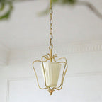 Unlit Clérachic pendant with a cream vertically pleated shade, hanging beside white wall panelling with a blurred plant behind.
