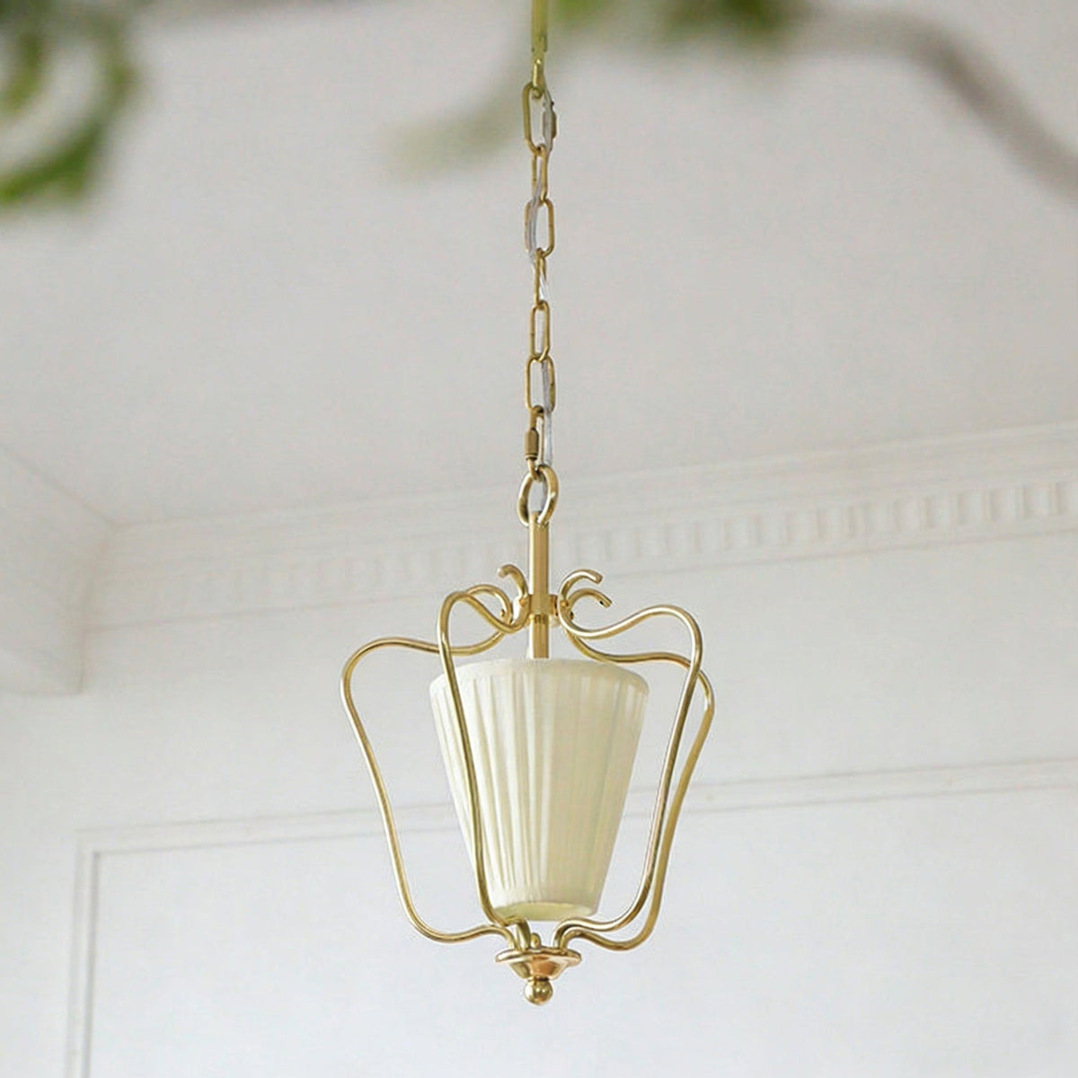 Unlit Clérachic pendant with a cream vertically pleated shade, hanging beside white wall panelling with a blurred plant behind.