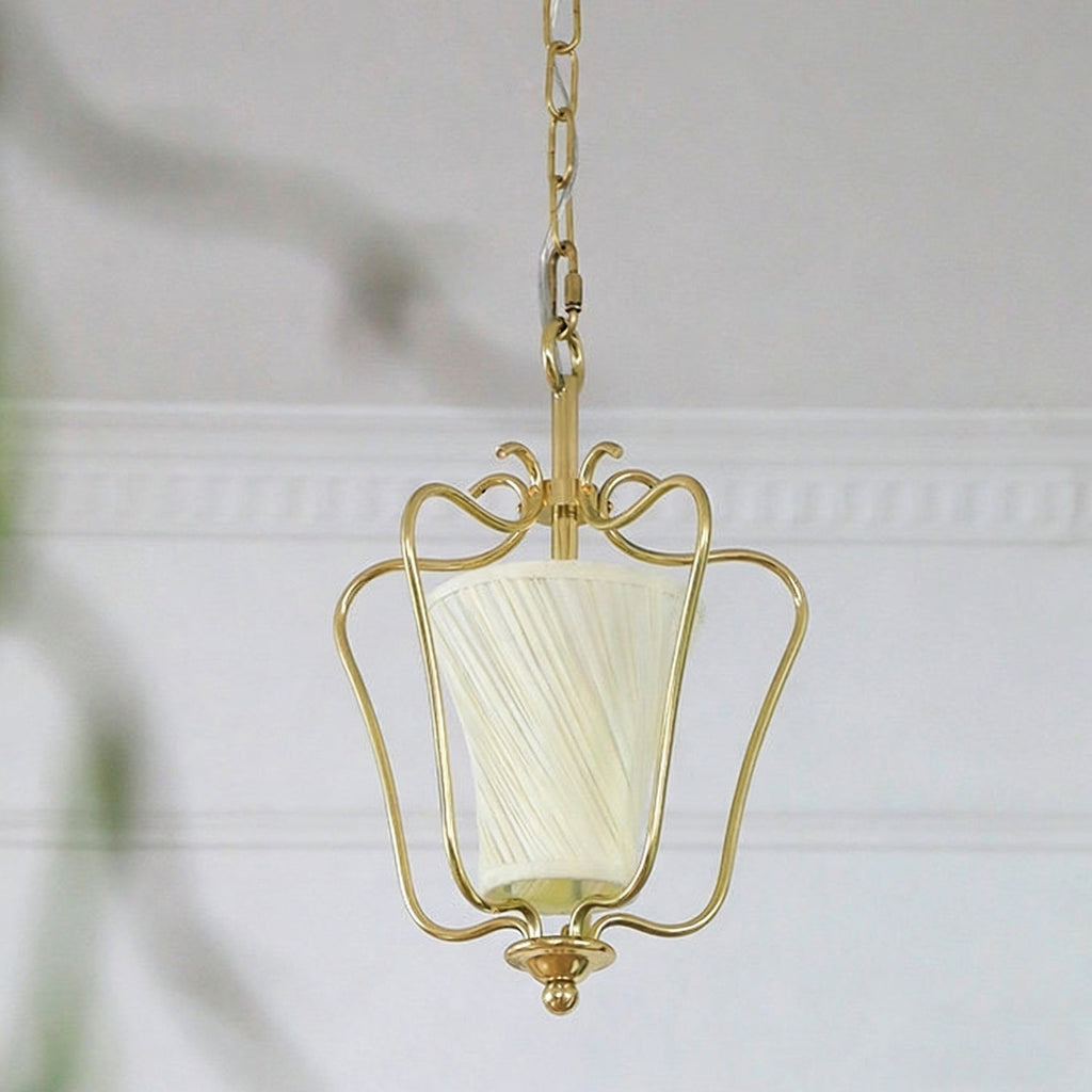 Unlit Clérachic brass pendant with a cream twisted-pleat shade, seen against white mouldings and a soft-focus leaf.