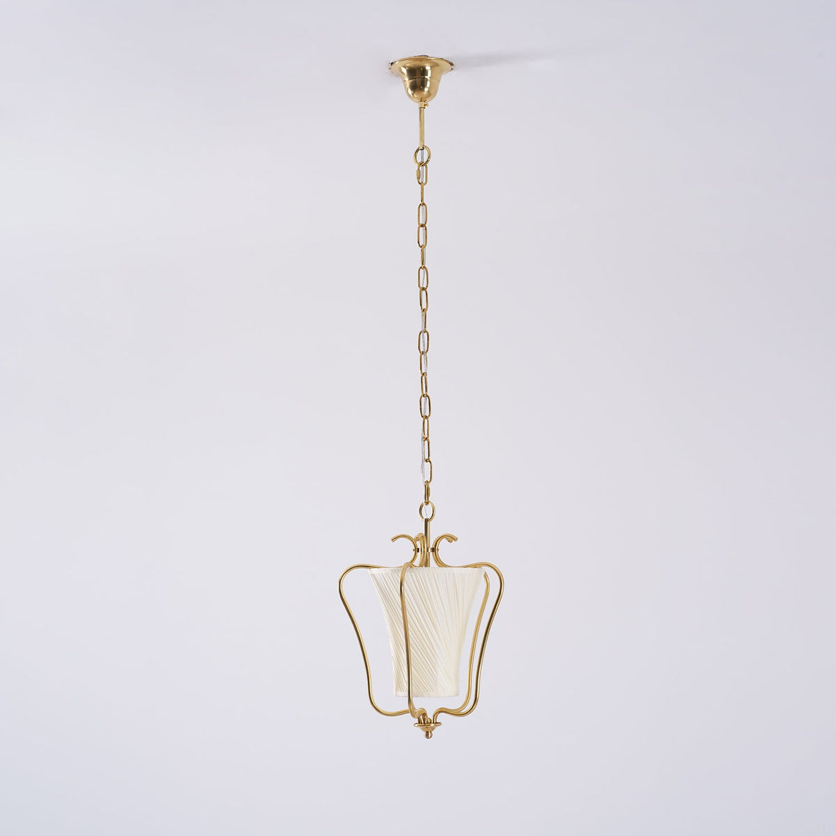 Clérachic brass pendant on a long chain, seen small against a pale wall to show how far it hangs.