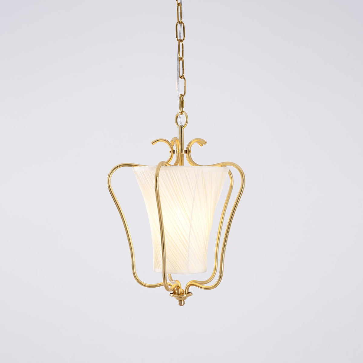Clérachic pendant with a lit diagonally pleated shade inside the brass frame, on a pale lilac-grey backdrop.
