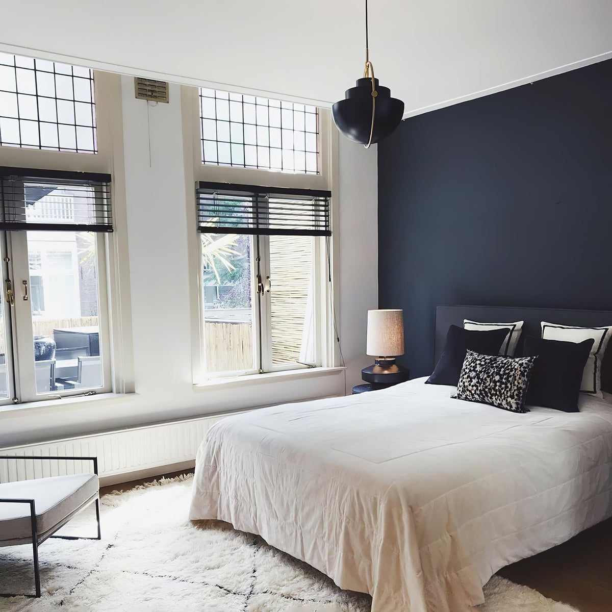 Black Clérachic pendant hung high in a bedroom with a navy wall, a white bed and tall windows.