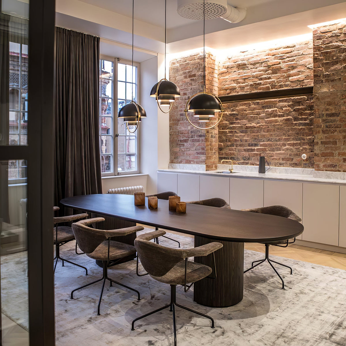 Three black Clérachic pendants with gold rings above a long dark oval table, in front of an exposed brick wall.
