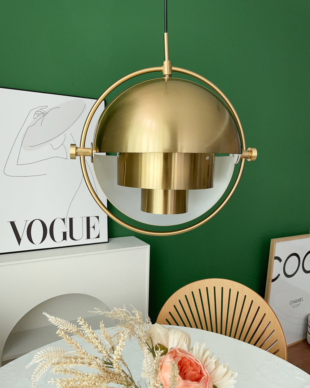 Gold-toned Clérachic pendant above a round table with a vase of dried flowers, against a green wall and a framed poster.
