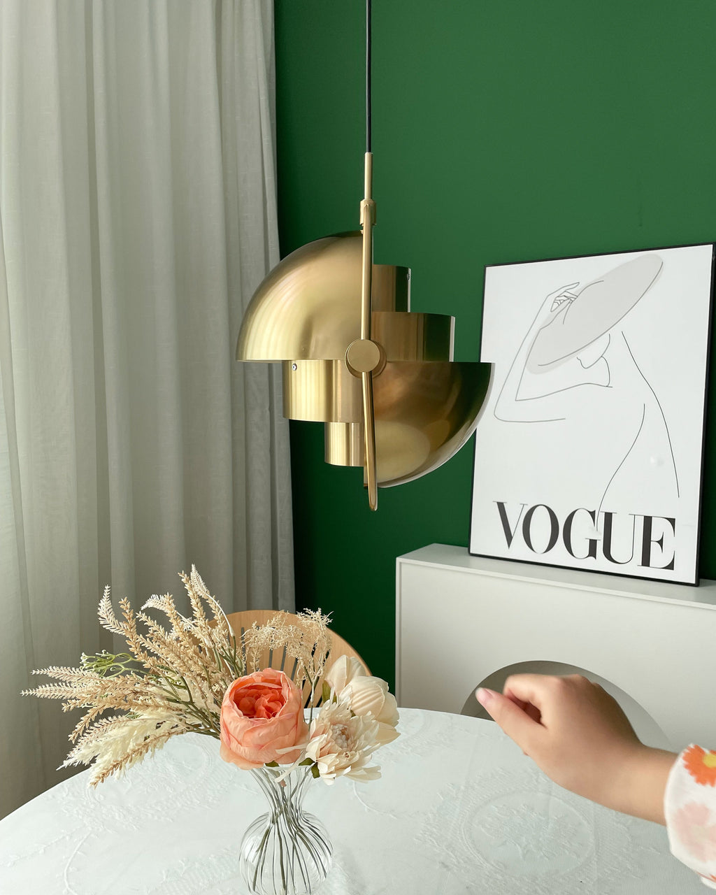 Gold-toned Clérachic pendant with its shade halves turned sideways, with a hand reaching in beside a green wall and a poster.