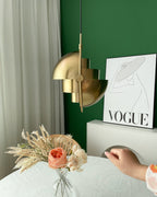 Gold-toned Clérachic pendant with its shade halves turned sideways, with a hand reaching in beside a green wall and a poster.