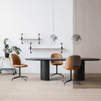 Two chrome Clérachic pendants over a dark oval table with tan chairs, against a white wall with shelves.