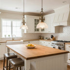 A pair of Clérachic pendants hangs above a butcher-block island with leather stools, in a white shaker-style kitchen.