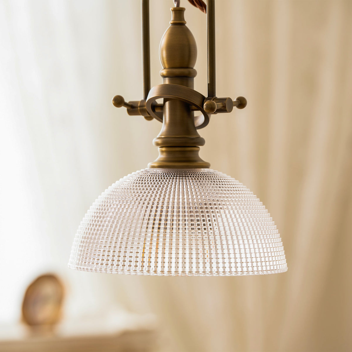 Close view of the ribbed glass dome and the forked antique brass top of this Clérachic pendant, with a cream curtain behind.